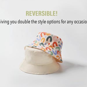 May include: A reversible bucket hat with a colorful abstract pattern on one side and a solid cream color on the other side. The hat is made of a lightweight fabric and has a wide brim.
