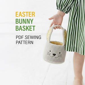 Peut inclure: Un panier de Pâques gris en forme de lapin, avec un visage souriant et de longues oreilles, est tenu par un enfant. L'intérieur du panier est jaune et il possède une anse. Le texte indique "EASTER BUNNY BASKET PDF SEWING PATTERN."