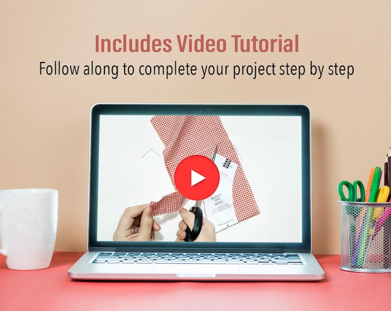 May include: A laptop computer displaying a video tutorial on how to cut fabric. The video shows a person's hands cutting a piece of red and white checkered fabric with a pair of scissors. The video player has a play button in the center of the screen. The laptop is on a pink surface with a white mug and a container of colorful pencils and scissors.