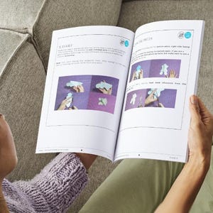 Peut inclure: Livre ouvert avec des instructions et des illustrations. Le livre est ouvert sur une page avec les titres "A DART" et "THE PIECES". Les illustrations montrent des instructions &eacute;tape par &eacute;tape. La personne porte un pull violet et est assise sur un canap&eacute; gris.