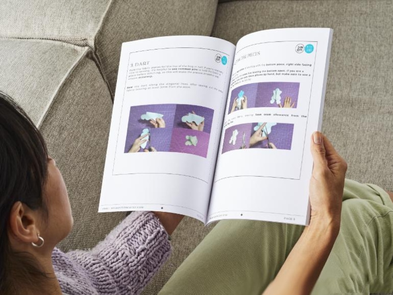Peut inclure: Livre ouvert avec des instructions et des illustrations. Le livre est ouvert sur une page avec les titres "A DART" et "THE PIECES". Les illustrations montrent des instructions &eacute;tape par &eacute;tape. La personne porte un pull violet et est assise sur un canap&eacute; gris.