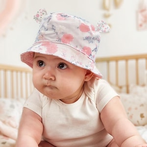 May include: A baby wearing a white and pink floral bucket hat with ear flaps. The hat has a pink brim and a white crown with pink flowers and green leaves.