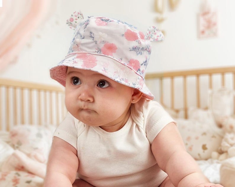 May include: A baby wearing a white and pink floral bucket hat with ear flaps. The hat has a pink brim and a white crown with pink flowers and green leaves.