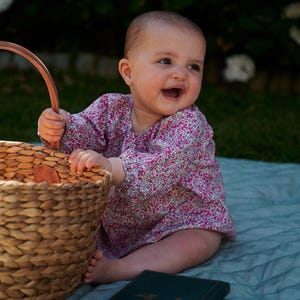 May include: A smiling baby wearing a pink floral print top sits on a blue blanket next to a woven basket and a book. The baby is holding the basket handle. The background features green grass and white flowers.
