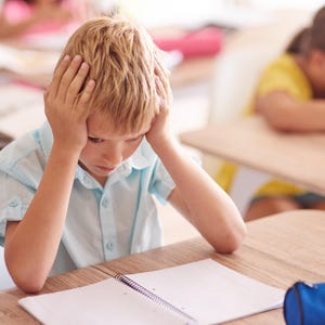 Puede incluir: Un niño pequeño de cabello rubio que viste una camisa de botones de manga corta de color azul claro, sentado en un escritorio de madera con las manos a los lados de la cabeza, mirando un cuaderno de espiral abierto con páginas blancas.