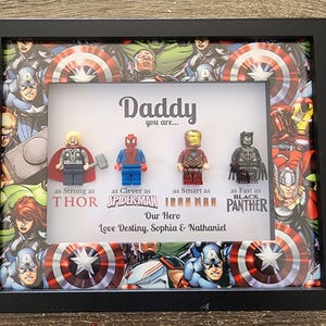 May include: A black shadow box frame containing four superhero minifigures arranged horizontally. The background features a comic book collage. Text reads: Daddy you are... as Strong as THOR, as Clever as SPIDER-MAN, as Smart as IRON MAN, as Fast as BLACK PANTHER. Our Hero, Love Destiny, Sophia & Nathaniel.