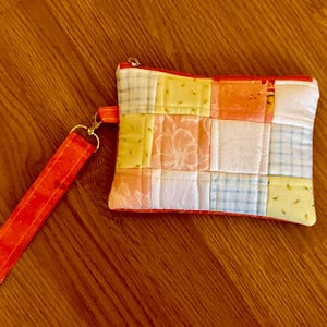 May include: A patchwork wristlet purse with a red wrist strap. The purse is made of small, square fabric patches in various pastel colors, including yellow, pink, and white. A gold zipper and red trim complete the design.