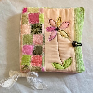 May include: A square fabric book cover featuring a patchwork panel of pink and green squares on the left and a solid peach panel on the right. A fabric flower appliqué with pink and yellow petals is stitched onto the peach section, secured by a black button.