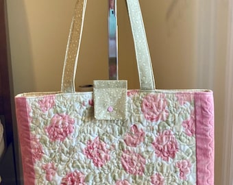 Puede incluir: Un bolso tote acolchado con un patrón floral en tonos rosa y verde. El bolso tiene un pequeño bolsillo en la parte delantera y dos asas.