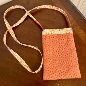 Puede incluir: Un pequeño bolso bandolera acolchado con una correa de estampado floral. El bolso está hecho de una tela de color melocotón con un estampado floral blanco. La correa está hecha de una tela blanca con un estampado floral de melocotón.