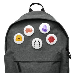 Könnte beinhalten: Ein anthrazitfarbener Rucksack mit fünf runden Ansteckbuttons. Die Buttons zeigen ein gelbes Umarmungs-Emoji, einen braunen, verpixelten Alien, einen violetten Kristall, die Umrisse eines schwarz-weißen Lamas und ein rundes rotes Wesen.
