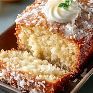 May include: A slice of coconut cake with a swirl of white frosting and a mint leaf garnish. The cake has a golden-brown crust and is topped with shredded coconut. The cake is on a brown rectangular plate.