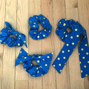 May include: A collection of blue scrunchies with gold polka dots. The set includes a scrunchie with a bow, a standard scrunchie, a scrunchie with a long tie, and a scrunchie with a large bow. The accessories are arranged on a wooden surface.