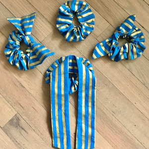 May include: A collection of blue and gold striped scrunchies and a matching hair scarf. The scrunchies are in various styles, including a bow and a classic circular design. The fabric has a shiny, metallic appearance.