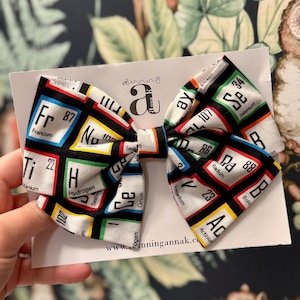 May include: A white bow tie featuring a black background with colorful periodic table elements. The bow tie has a central knot with a repeating pattern of chemical symbols and atomic numbers. The text "dinningannak.com" is visible on the card.