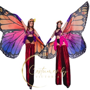 Puede incluir: Dos artistas con alas de mariposa y zancos de terciopelo rojo. Las alas son multicolores con tonos de naranja, rosa, morado y azul. Las artistas llevan coronas de flores y tops de lentejuelas.