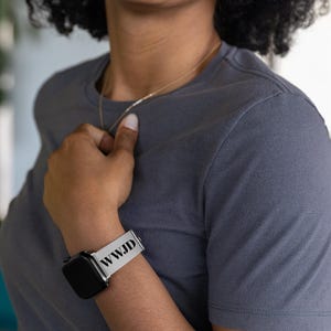 May include: A black smartwatch with a silver band that reads "WWJD" in black text. The watch is worn on a person's wrist. The person is wearing a gray t-shirt and a gold necklace.