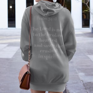 May include: A grey hoodie featuring the text "Psalm 34:18" and a religious quote on the back. The hoodie has a hood and long sleeves. A brown handbag with a chain strap hangs on the side.