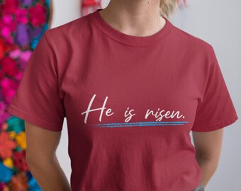Könnte beinhalten: Ein weinrotes T-Shirt mit dem Text "He is risen" in weißer Schrift, unterstrichen mit einer blauen Linie. Das T-Shirt hat einen Rundhalsausschnitt.