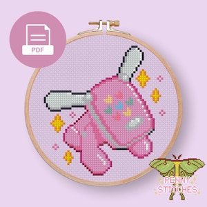 May include: A cross-stitch pattern in a wooden embroidery hoop featuring a pink, blocky, rabbit-like character with long ears and small colorful butterfly motifs. The design is set against a light purple background with yellow four-pointed stars.