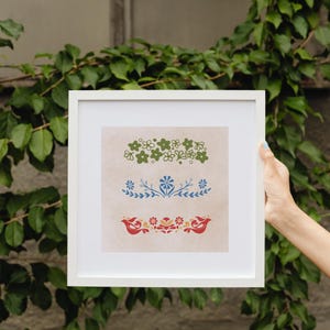 May include: A white frame with a beige background featuring three floral designs. The top design is a cluster of green flowers. The middle design is a blue floral sprig. The bottom design is a red and orange floral design with two birds.