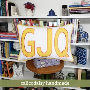 May include: A white pillow with a red border and three large yellow letters, "GJQ", sewn on the front. The pillow is sitting on a wooden table in front of a bookshelf.
