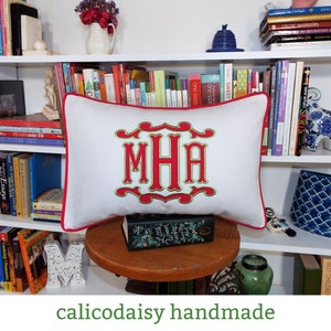 May include: White pillow with red trim and a green and red embroidered monogram "MHA". The pillow is sitting on a wooden table with a book on top.