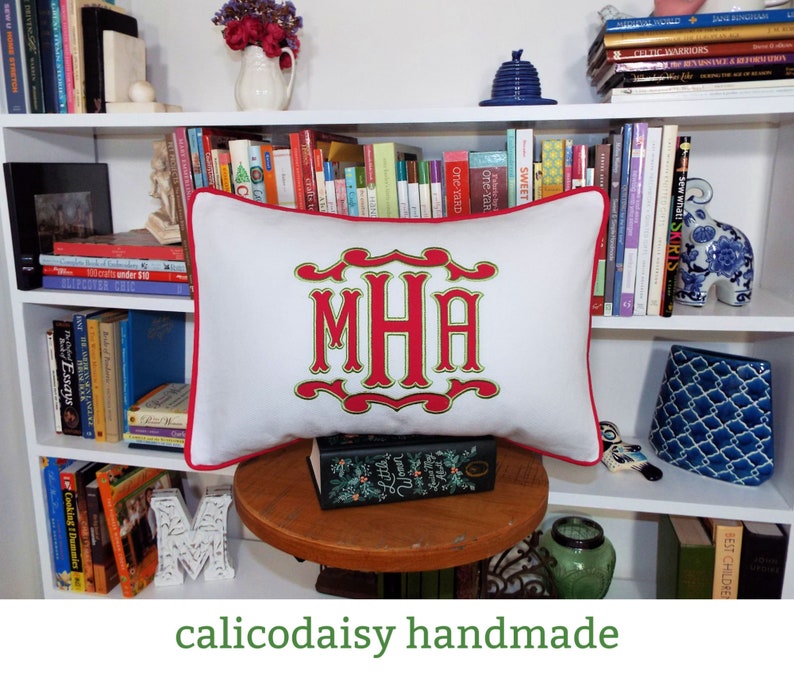 May include: White pillow with red trim and a green and red embroidered monogram "MHA". The pillow is sitting on a wooden table with a book on top.