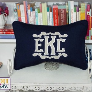 May include: A navy blue pillow with a beige monogram of the letters "EKC" in a decorative font. The pillow is sitting on a white shelf with books behind it.