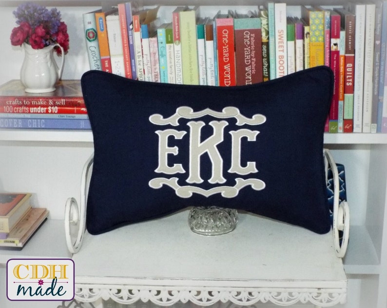 May include: A navy blue pillow with a beige monogram of the letters "EKC" in a decorative font. The pillow is sitting on a white shelf with books behind it.