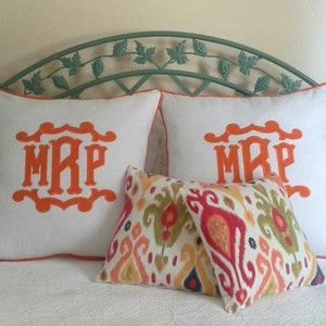 May include: Two white pillowcases with orange embroidered initials "MRP" and a colorful ikat patterned throw pillow on a bed with a green metal headboard.
