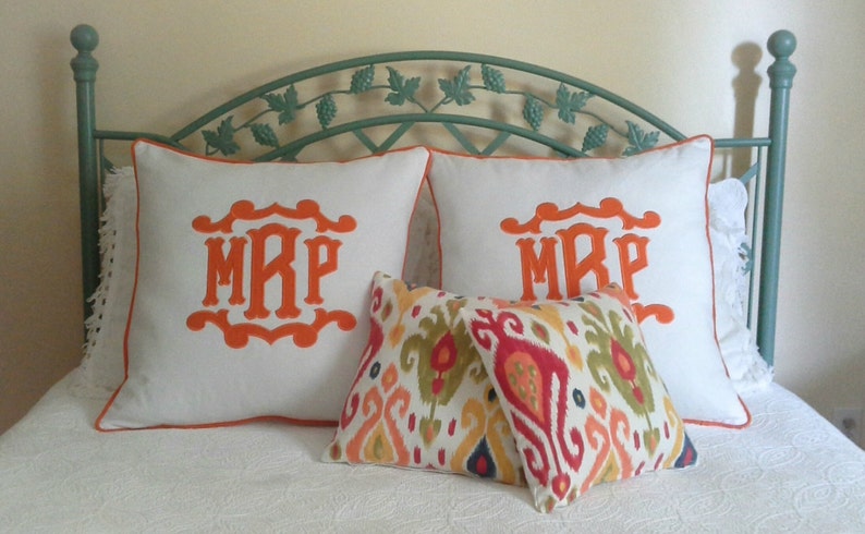 May include: Two white pillowcases with orange embroidered initials "MRP" and a colorful ikat patterned throw pillow on a bed with a green metal headboard.