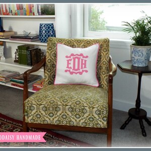 May include: A green and gold patterned armchair with a white pillow featuring a pink monogram "EDH". The chair is in a room with a bookshelf and a small table with a potted plant.