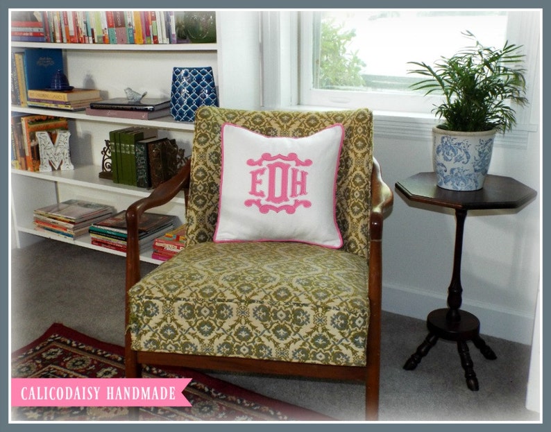 May include: A green and gold patterned armchair with a white pillow featuring a pink monogram "EDH". The chair is in a room with a bookshelf and a small table with a potted plant.