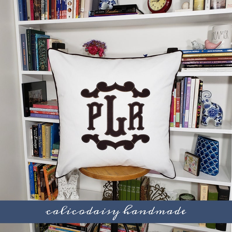 May include: White pillow with a brown monogram of the letters "PLR" in a decorative frame. The pillow is sitting on a wooden stool in front of a white bookshelf filled with books.