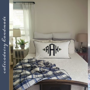May include: A white bed with a blue and white plaid throw blanket. The bed has a white pillow with black embroidery that says "AAK".