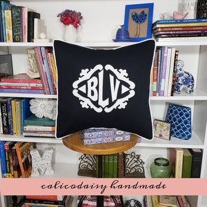 May include: A black and white monogram pillow with the letters "BLV" embroidered in white on a black background. The pillow is sitting on a wooden shelf with books and other decorative items.