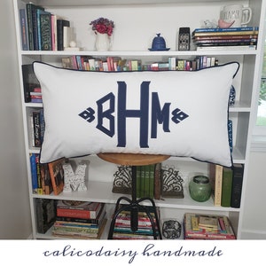 May include: A white pillow with a navy blue monogram "BHM" on a white bookshelf with books. The pillow has a navy blue trim.