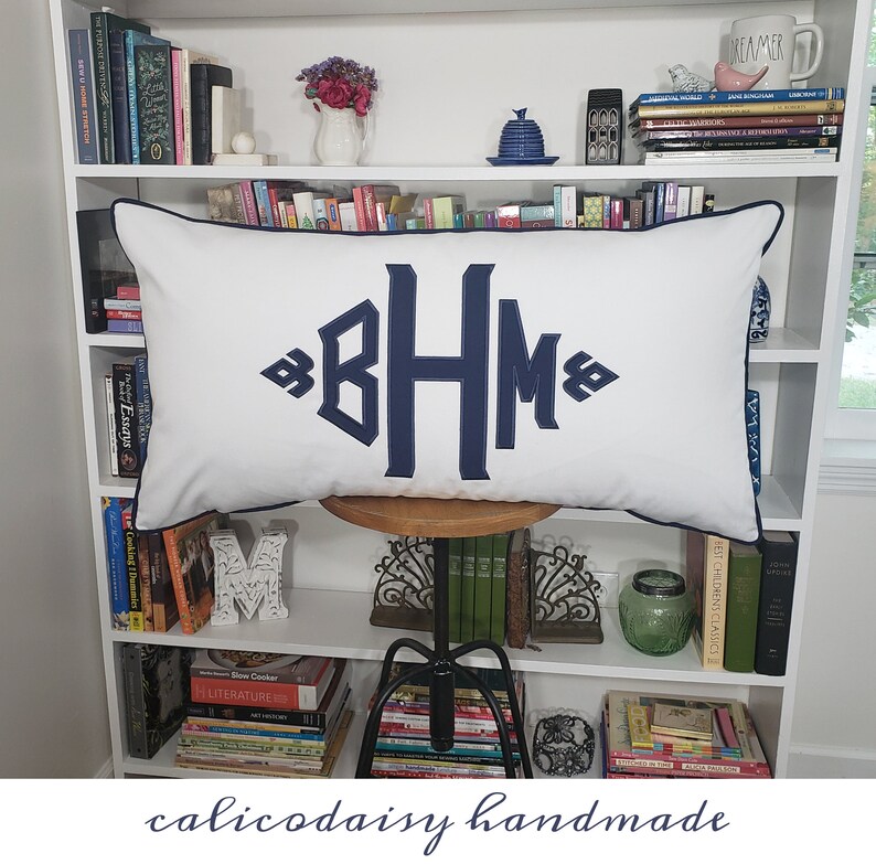 May include: A white pillow with a navy blue monogram "BHM" on a white bookshelf with books. The pillow has a navy blue trim.