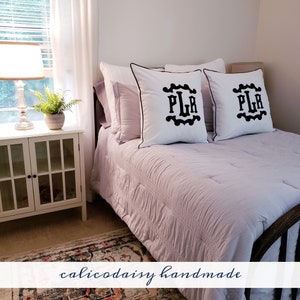 May include: Two white pillowcases with black trim and black embroidered initials "PLR" in a decorative font. The pillows are on a light purple bedspread with a white quilted pattern.