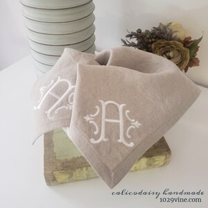 May include: Two beige linen napkins with white embroidered letter "A" monograms. The napkins are folded and stacked on top of a vintage book.
