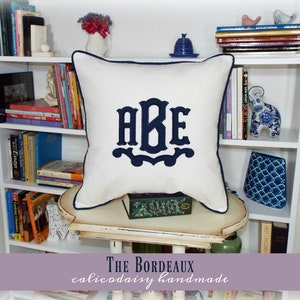 May include: White pillow with navy blue trim and a navy blue monogram of the letters "ABE" in a decorative font. The pillow is sitting on a white shelf with books and decorative items.