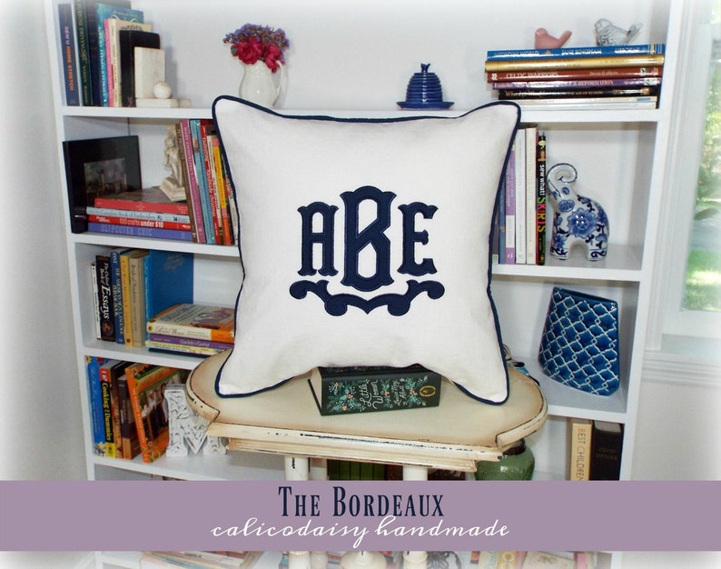 May include: White pillow with navy blue trim and a navy blue monogram of the letters "ABE" in a decorative font. The pillow is sitting on a white shelf with books and decorative items.