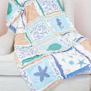 May include: A patchwork rag quilt featuring square fabric panels in shades of blue, teal, and tan. The quilt displays various nautical patterns including starfish, seahorses, and seashells, with frayed, exposed seams between each square block.