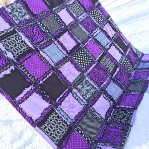 May include: A handmade patchwork quilt featuring a variety of fabrics in shades of purple, black, and white. The quilt has a grid pattern with squares of solid purple, polka dots, zebra print, and geometric designs. The quilt is displayed on a snowy surface.