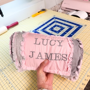 May include: A pink and gray decorative pillow with the name "LUCY JAMES" embroidered in gray thread. The pillow has a ruffled edge and is held by a person.