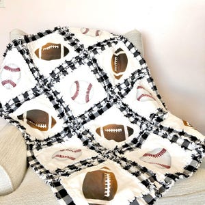 May include: A rag quilt featuring a repeating pattern of white squares printed with brown footballs and white baseballs with red stitching. The squares are separated by black and white buffalo plaid fabric strips with frayed, exposed seams.