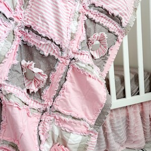May include: A handmade baby quilt in shades of pink, gray, and white. The quilt features a patchwork design with ruffled edges and decorative fabric flowers. The quilt is next to a white crib.
