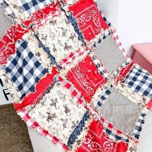 May include: A patchwork quilt with a Western theme, featuring squares of red bandanas, navy and white gingham, and cowboy-themed fabric. The quilt has a ruffled edge and is draped over a light gray chair. The overall color scheme is red, white, and blue.
