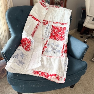 May include: A white and red patchwork quilt with a floral pattern, draped over a blue armchair. The quilt features a mix of textures and patterns, with red and blue accents. The chair has a button-tufted back.
